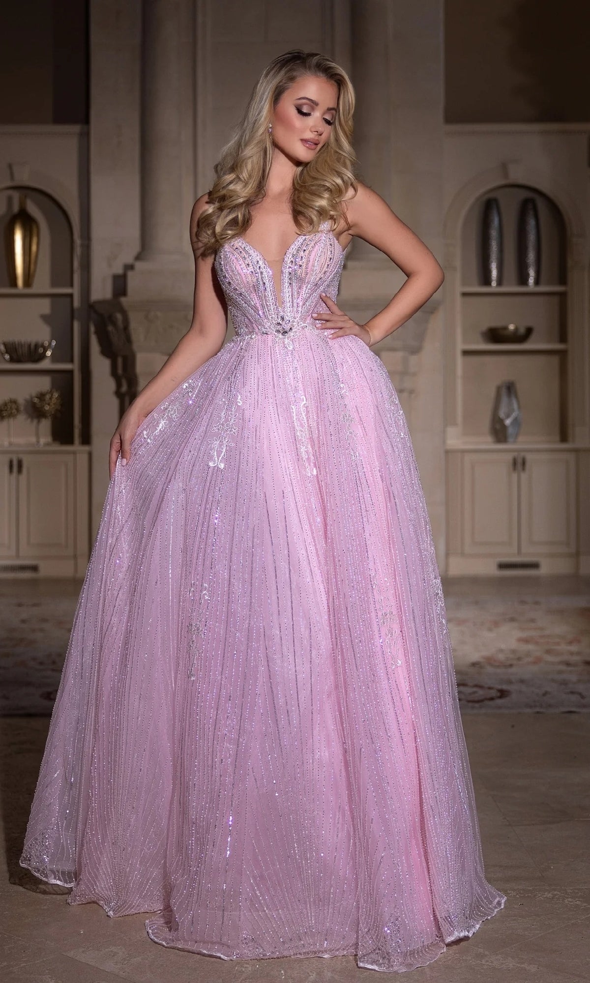 A woman in the Portia and Scarlett PS25310 long prom dress, a floor-length light pink ball gown with sequin embellishments, stands indoors before built-in shelves.